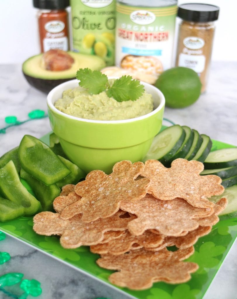 Avocado Hummus in bowl plated with shamrock-shaped tortilla chips, sliced cucumber, and green bell pepper